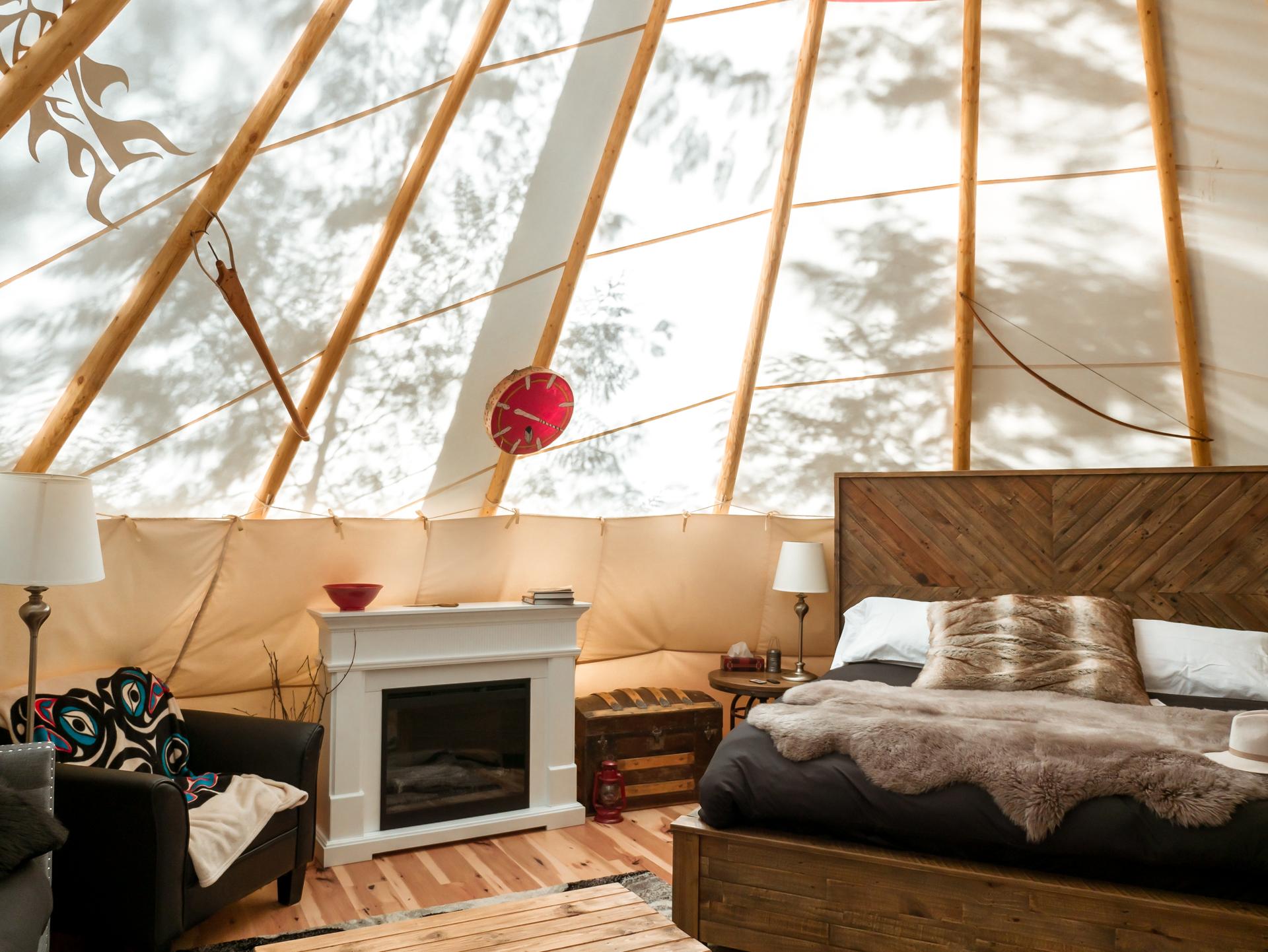 Interior of a luxury teepee in British Columbia with a made bed, fireplace, and Indigenous artwork beneath exposed lodgepole framing