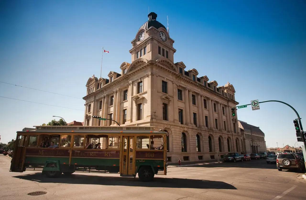 A vintage green and gold Moose Jaw Electric Railway trolley car numbered 100 passing in front of the ornate Beaux-Arts style Moose Jaw City Hall clock tower flying a Canadian flag in Moose Jaw, Saskatchewan, Canada on a clear blue sky day