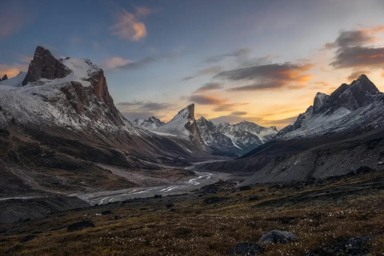 Dramatic snow-capped peaks including Mount Thor rising above a wide glacial valley with a winding braided river on Baffin Island, Nunavut, Canada, under a vivid orange and blue sunset sky with scattered clouds