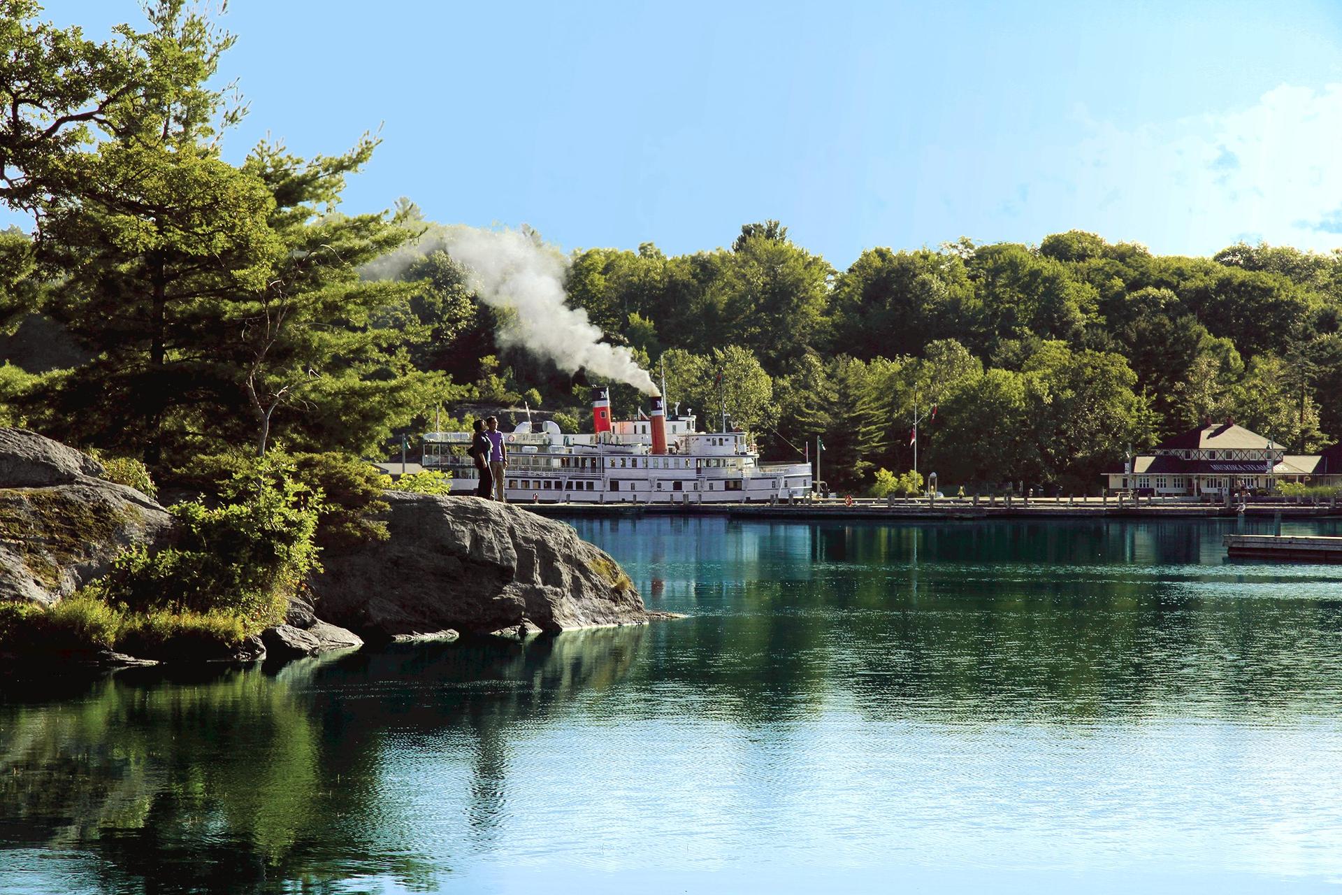 A couple watches the Muskoka Steamships vessel depart from a granite outcrop on Lake Muskoka, Ontario, cottage country