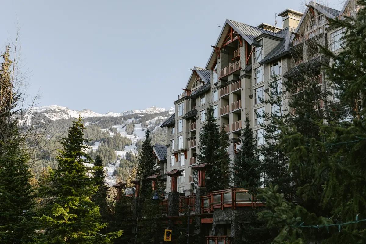 Tall evergreen trees frame the Pan Pacific Whistler Village Centre hotel, with snow-dusted mountain ski runs in the background.