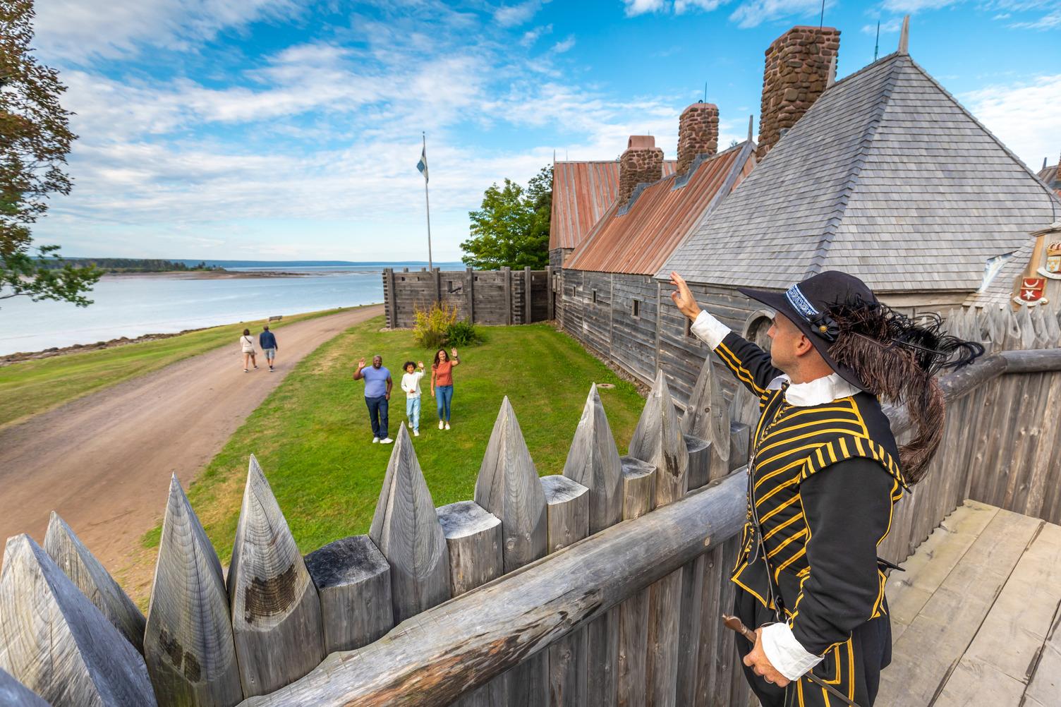 A costumed interpreter in 17th-century dress stands on the palisade at Port-Royal National Historic Site, gesturing toward the Habitation as a family waves from the grounds below.