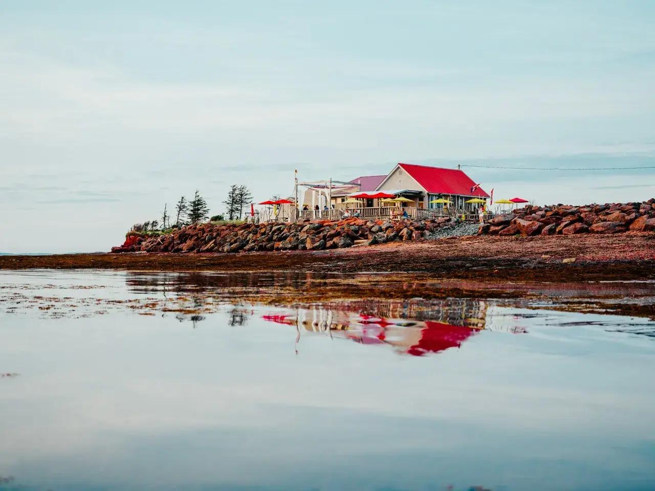 A coastal building with a bright red roof and colourful outdoor umbrellas perched on a rocky red sandstone shoreline in Prince Edward Island, Canada, with its mirror-like reflection visible in the shallow tidal water in the foreground under an overcast sky