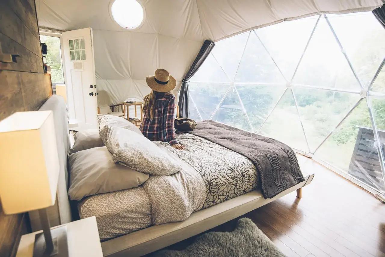 A woman wearing a straw hat and plaid shirt sitting on a neatly made queen bed inside a geodesic glamping dome at Ridgeback Lodge in New Brunswick, Canada, gazing out through the large transparent triangular window panels at a lush green forest
