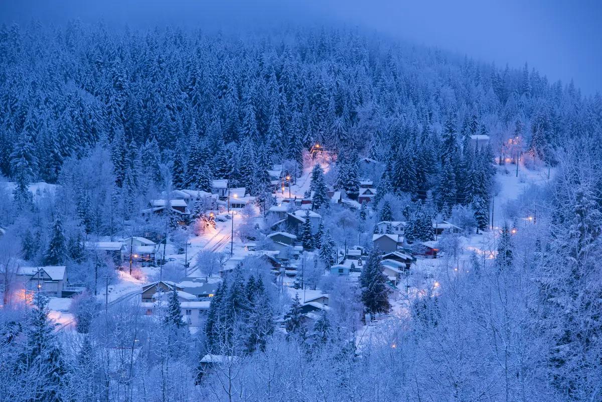 Warm streetlights glow through snow-covered homes and frosted evergreens blanketing the hillside town of Rossland, BC at dusk.
