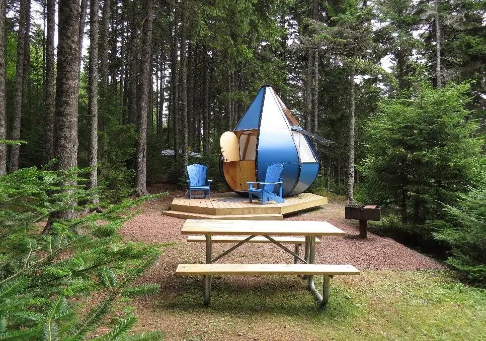 A blue teardrop-shaped glamping cabin nestled in the forest at Fundy National Park, New Brunswick, with Adirondack chairs outside.