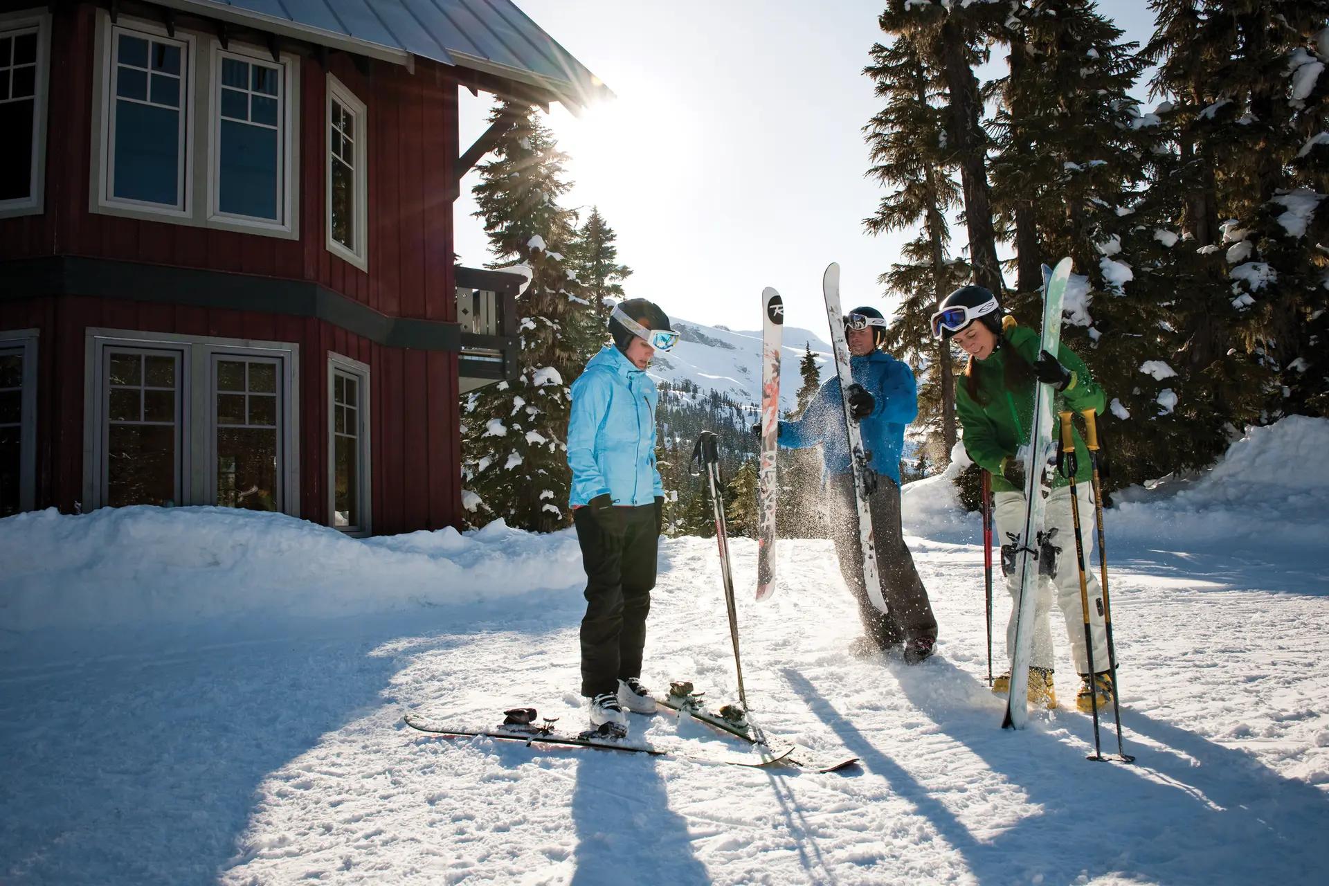 Three skiers in colorful gear stand outside a red ski-in/ski-out lodge at Whistler Blackcomb on a sunny day.