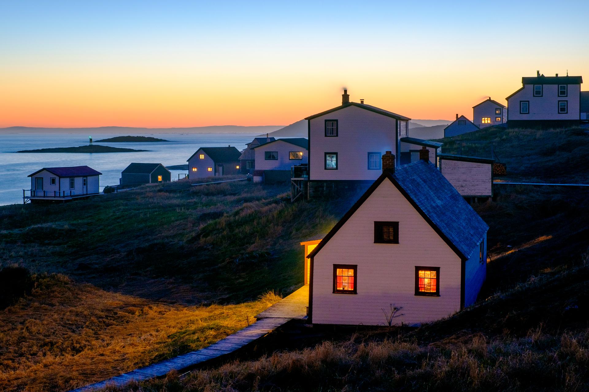Warmly lit wooden heritage houses connected by boardwalks scattered across a grassy slope above the water at sunset in the restored outport of Battle Harbour, Labrador