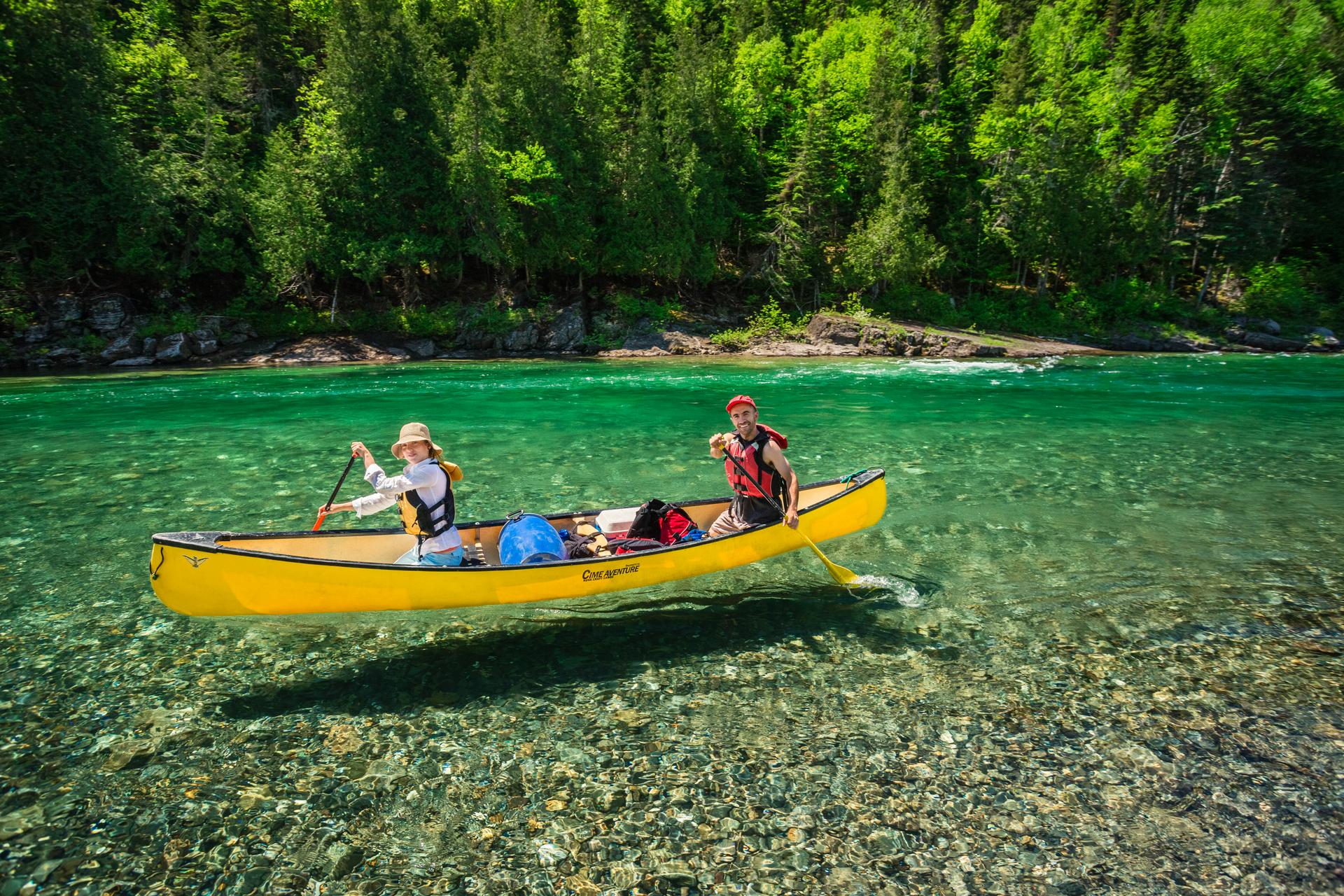 Bonaventure River in Gaspésie, where two paddlers in a yellow Cime Aventure canoe glide over water so clear the rocky riverbed is visible beneath them.