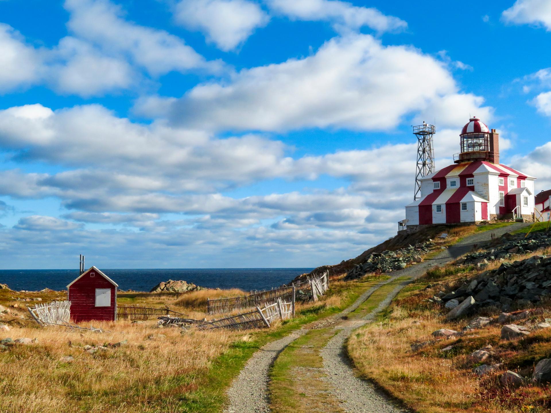 Red-and-white striped Cape Bonavista Lighthouse on a rocky headland above the sea, with a gravel path and a small red shed under a bright cloudy sky, Bonavista Peninsula, Newfoundland
