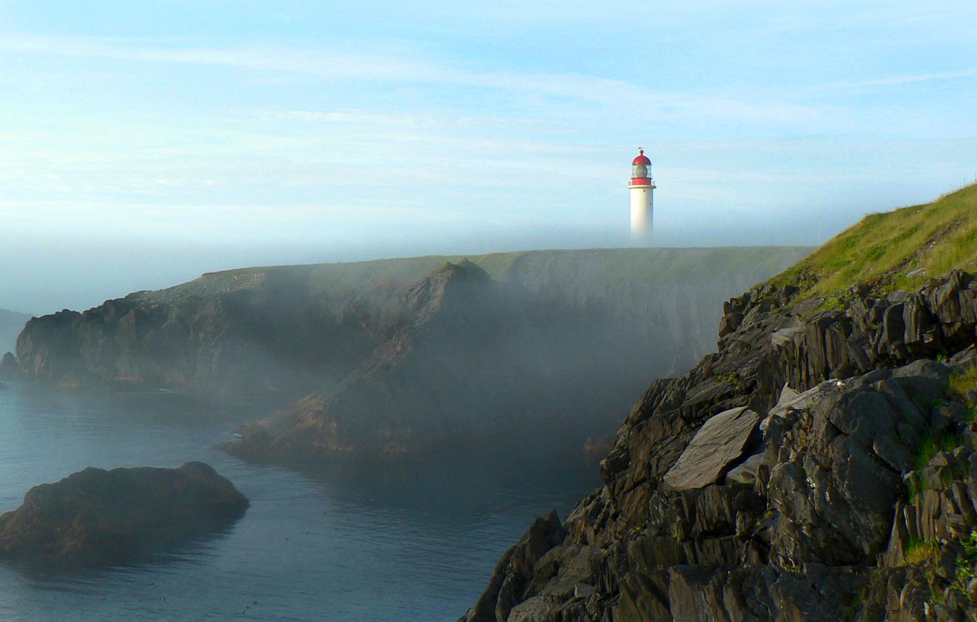 White and red Cape Race lighthouse rising above fog-shrouded coastal cliffs and dark rocky shoreline on the Avalon Peninsula, Newfoundland