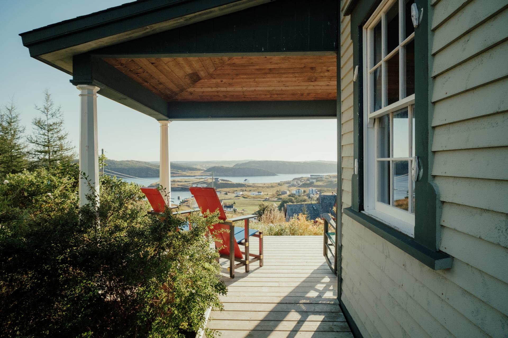 Covered wooden porch with red Adirondack chairs overlooking a coastal village and harbour in the evening light at Port Rexton, Newfoundland