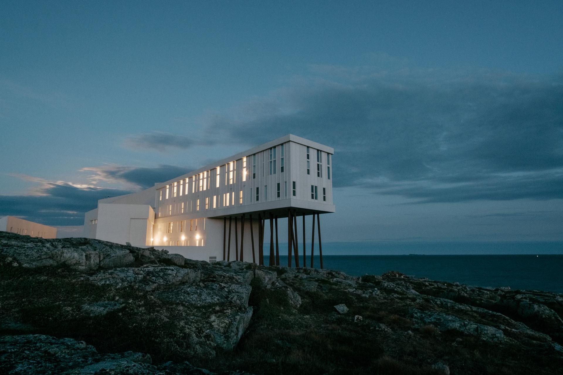 The white, angular Fogo Island Inn glowing at dusk, its long form raised on stilts over the rocky Fogo Island shoreline with the Atlantic Ocean stretching out behind it