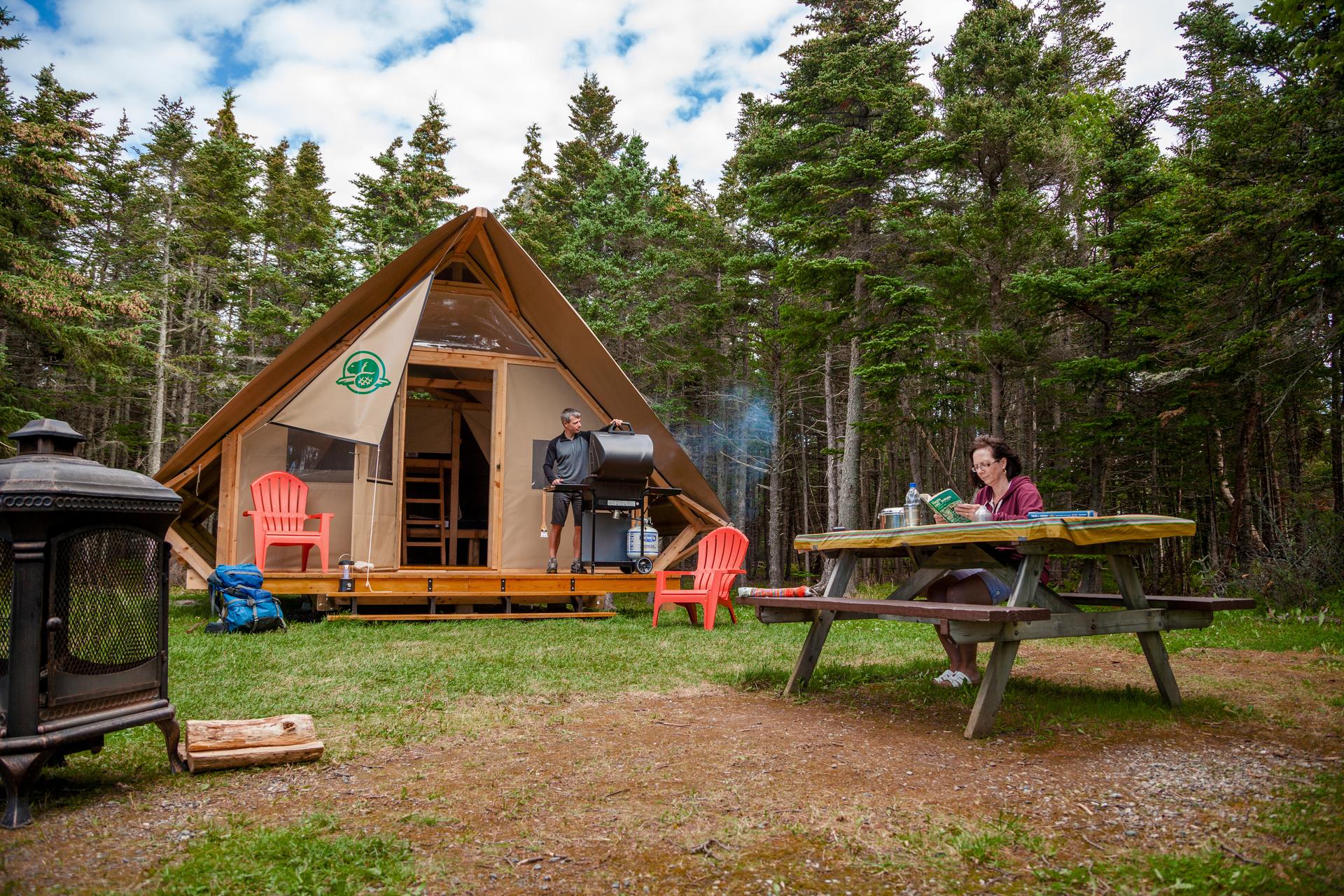Two people relaxing at their campsite in Gros Morne National Park, one grilling on the wooden deck of a canvas-and-wood oTENTik tent and the other reading at a picnic table, surrounded by boreal forest