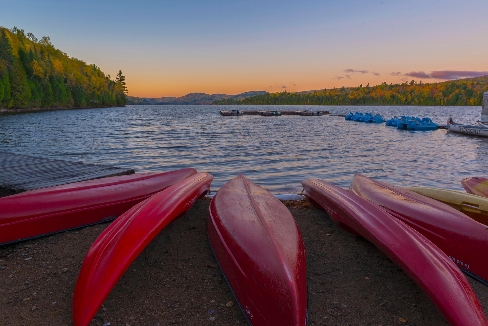 Red canoes rest upturned on a sandy shore at sunset in La Mauricie National Park, facing a calm lake with a dock, moored pedal boats, and forested hills in fall colour across the water.