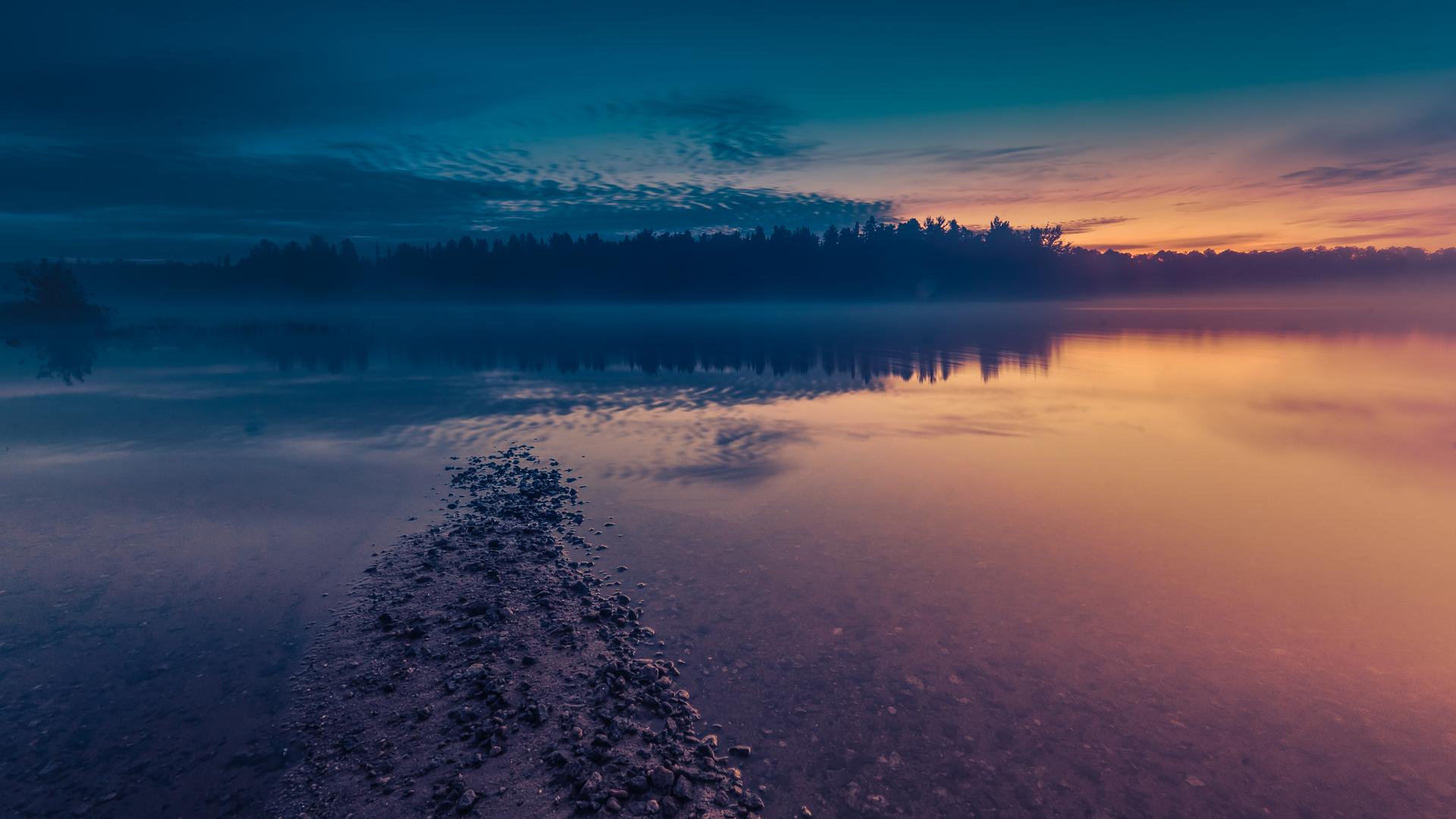 A calm lake at La Vérendrye Wildlife Reserve at sunset, with mist over the water, a rocky spit in the foreground, and a silhouetted treeline reflecting orange and blue.