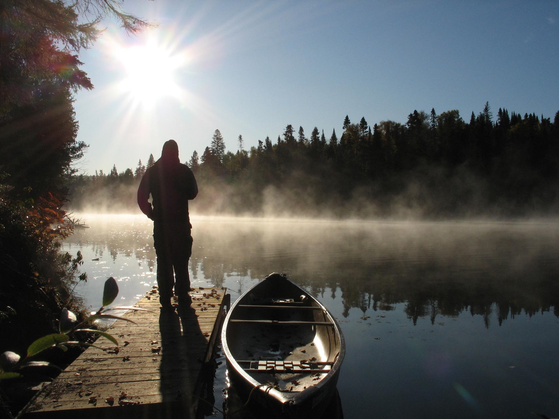 A person stands silhouetted on a wooden dock at sunrise in Parc national du Mont-Tremblant, beside a moored canoe, as mist rises off a still lake ringed by dark evergreen forest.