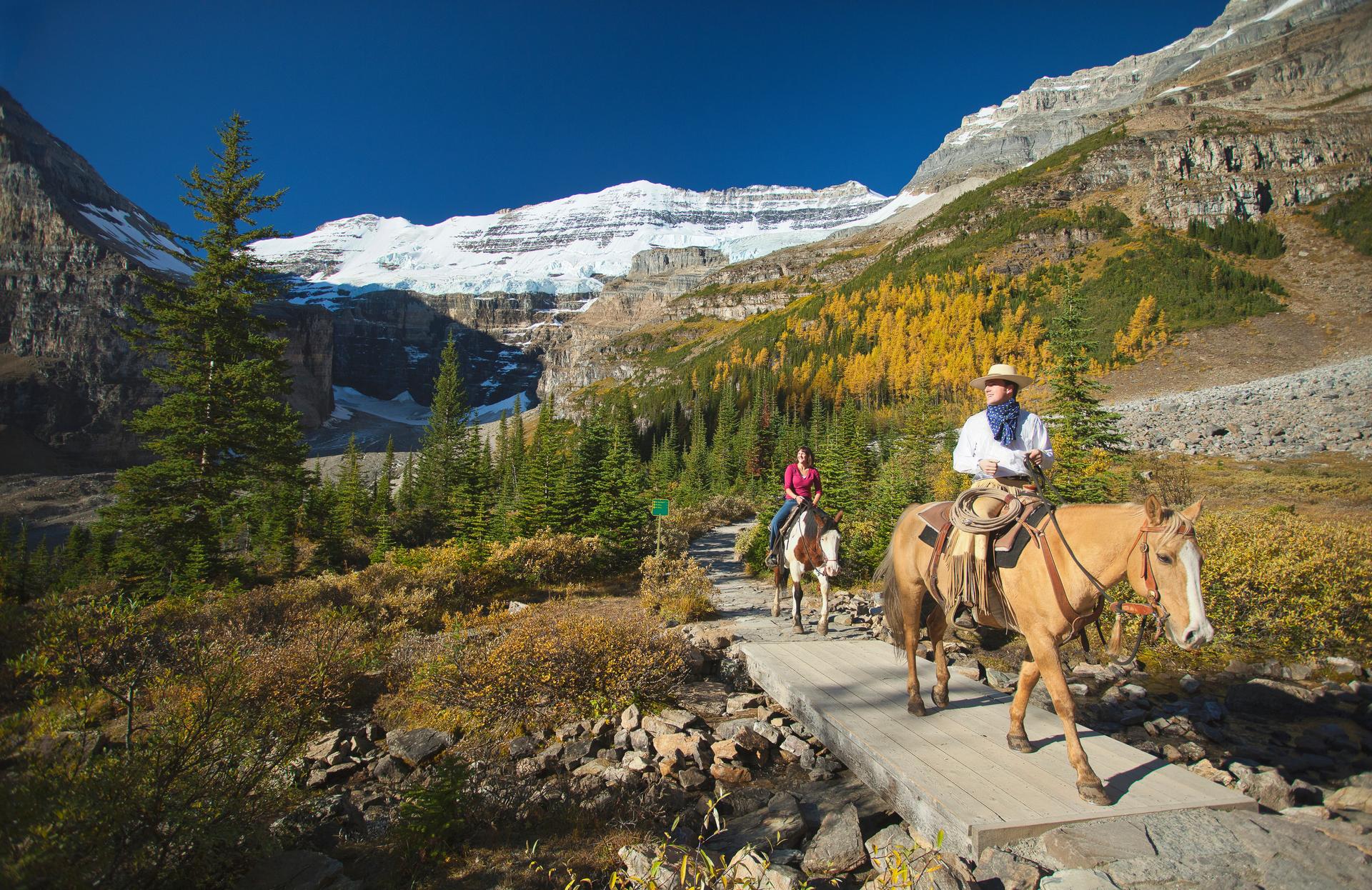 Horseback riders cross a wooden bridge on the trail to Plain of Six Glaciers, with glaciers and golden larch trees in the background near Lake Louise