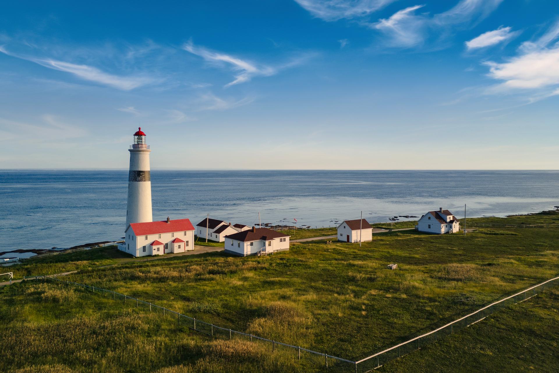 The tall white Point Amour Lighthouse with a red lantern room, surrounded by red-roofed heritage buildings on a green grassy point above the calm Atlantic in southern Labrador
