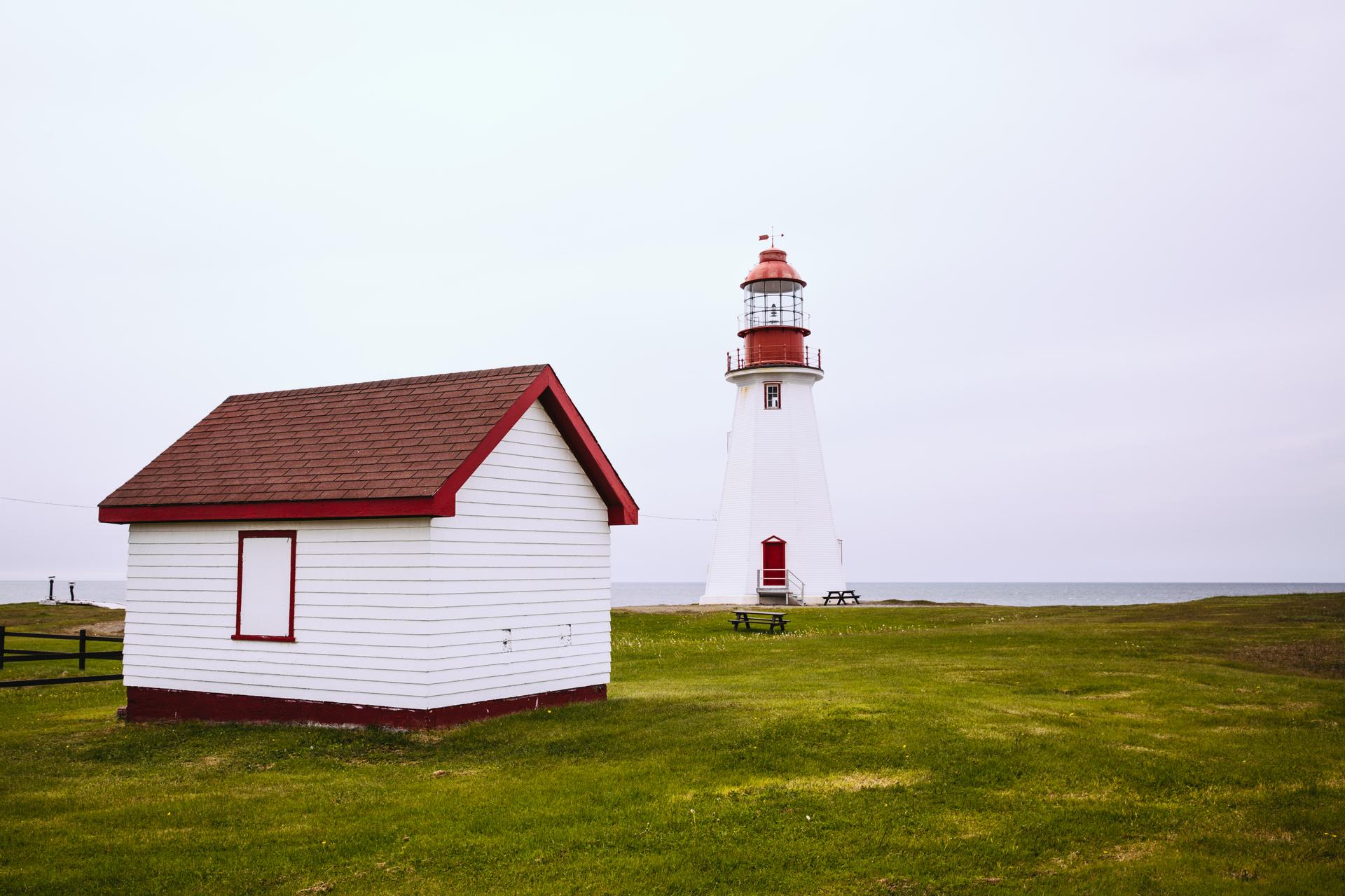 A white lighthouse with a red lantern room and a matching red-roofed outbuilding standing on a green grassy point above the Atlantic under an overcast sky