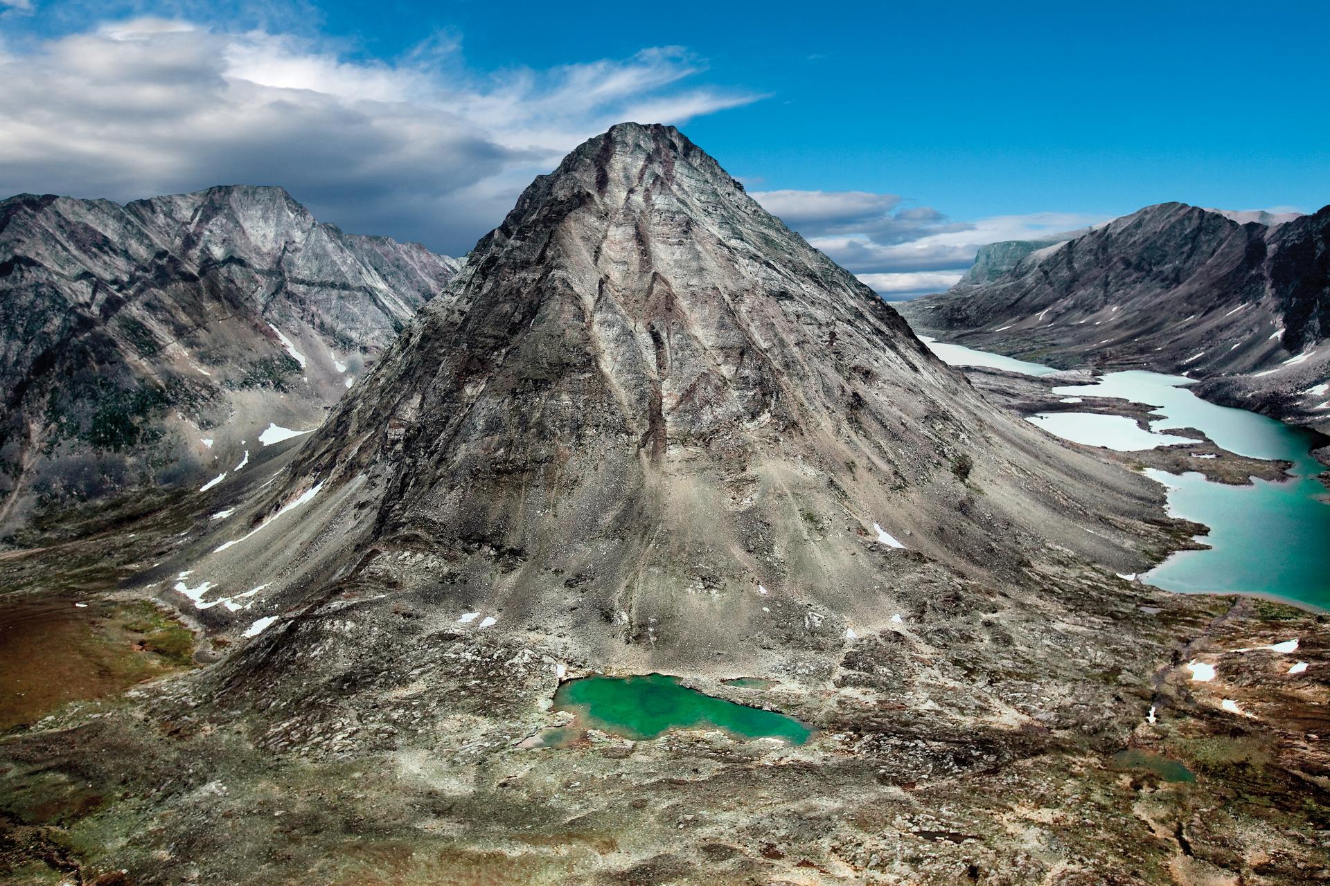 Aerial view of the rugged grey peaks of the Torngat Mountains in northern Labrador, with glacial turquoise lakes winding through the roadless wilderness below a blue sky with scattered clouds