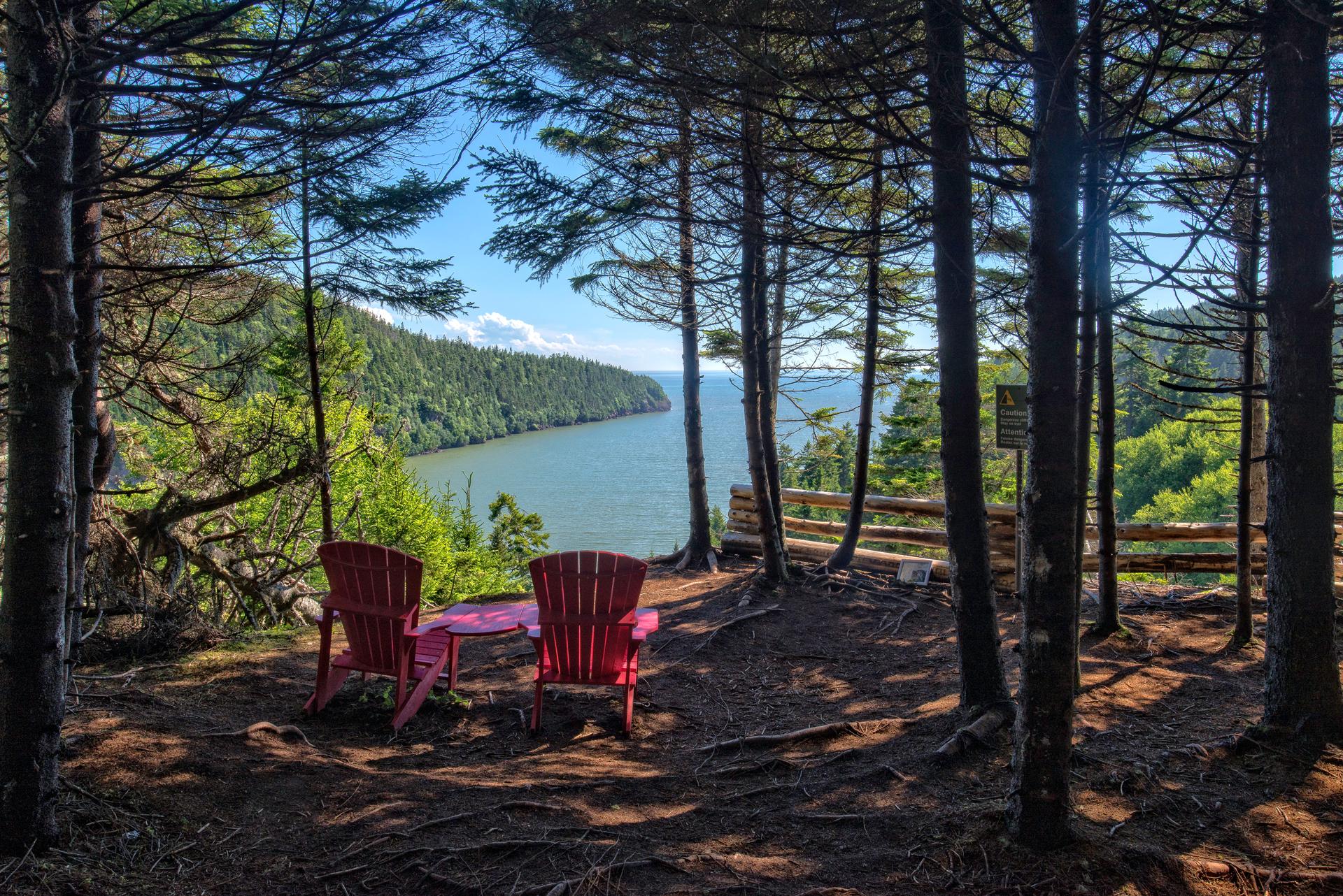 Two red Adirondack chairs at a forest lookout in Fundy National Park, New Brunswick, framed by Acadian forest above the Bay of Fundy.