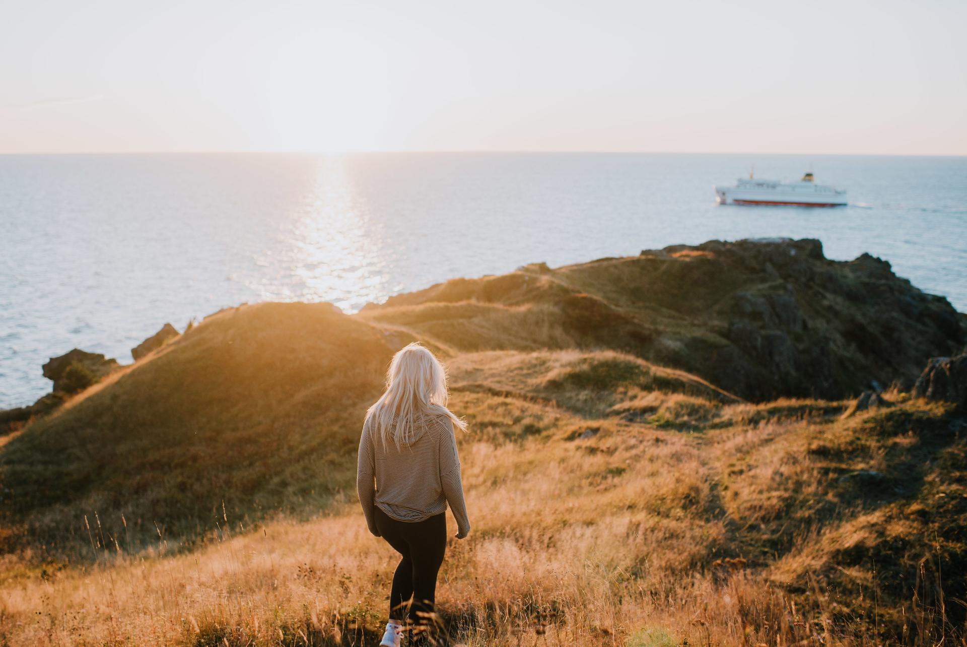 A hiker on a grassy clifftop at sunset on Grand Manan Island, New Brunswick, watching a ferry cross the Bay of Fundy.