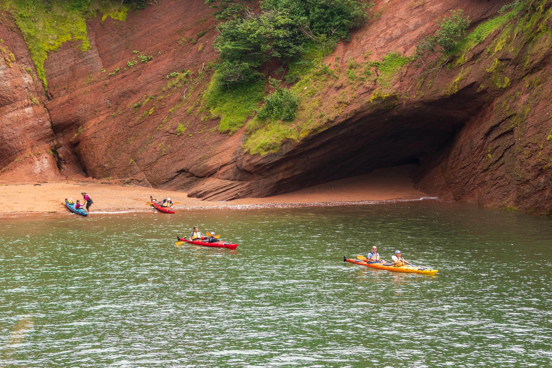 Kayakers paddling past the red sandstone St. Martins Sea Caves in New Brunswick, with a sandy beach exposed at the cave mouth.