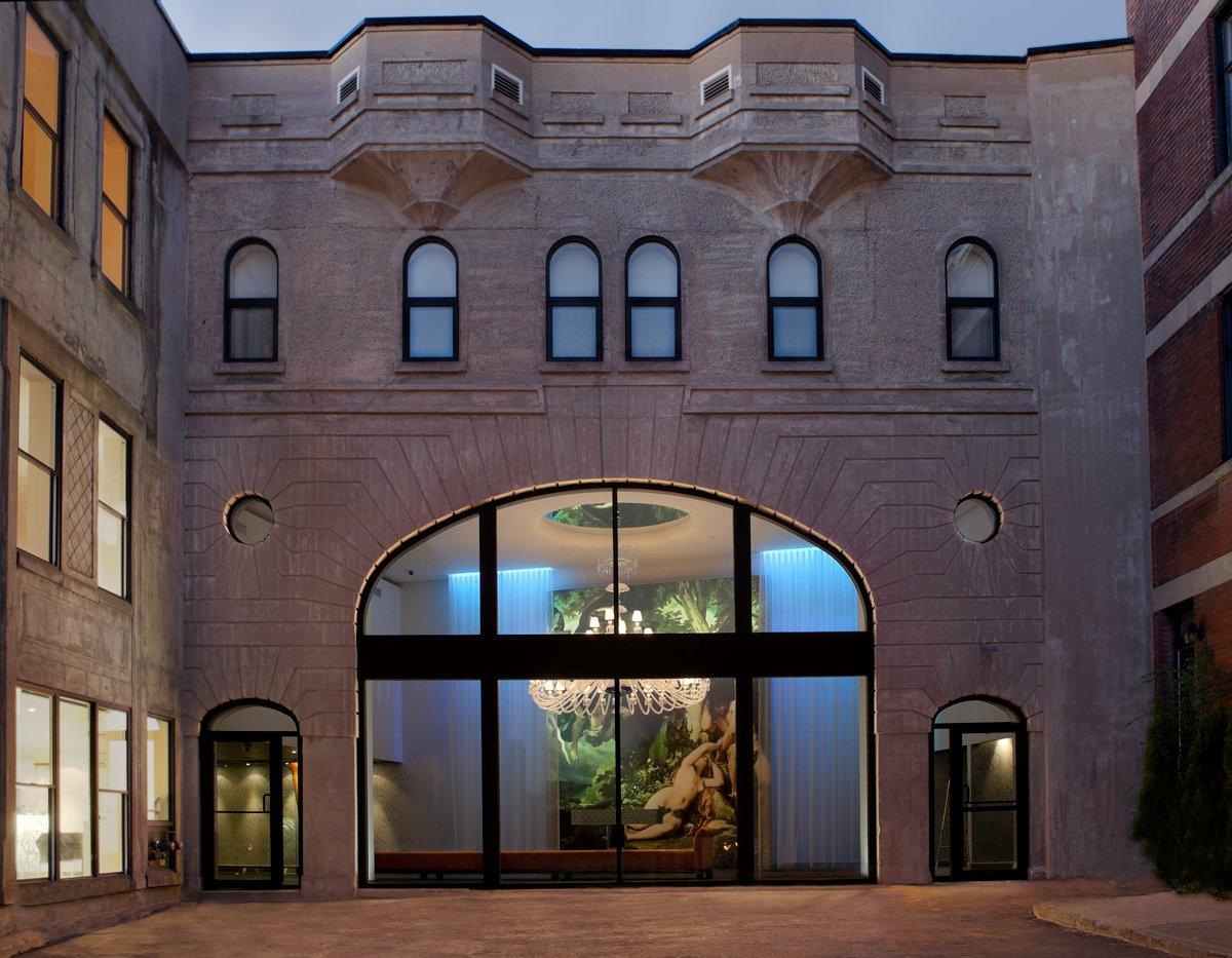 Illuminated stone facade and glass-arched entrance of the Loft Hotel Montréal with a large mural and chandelier visible inside