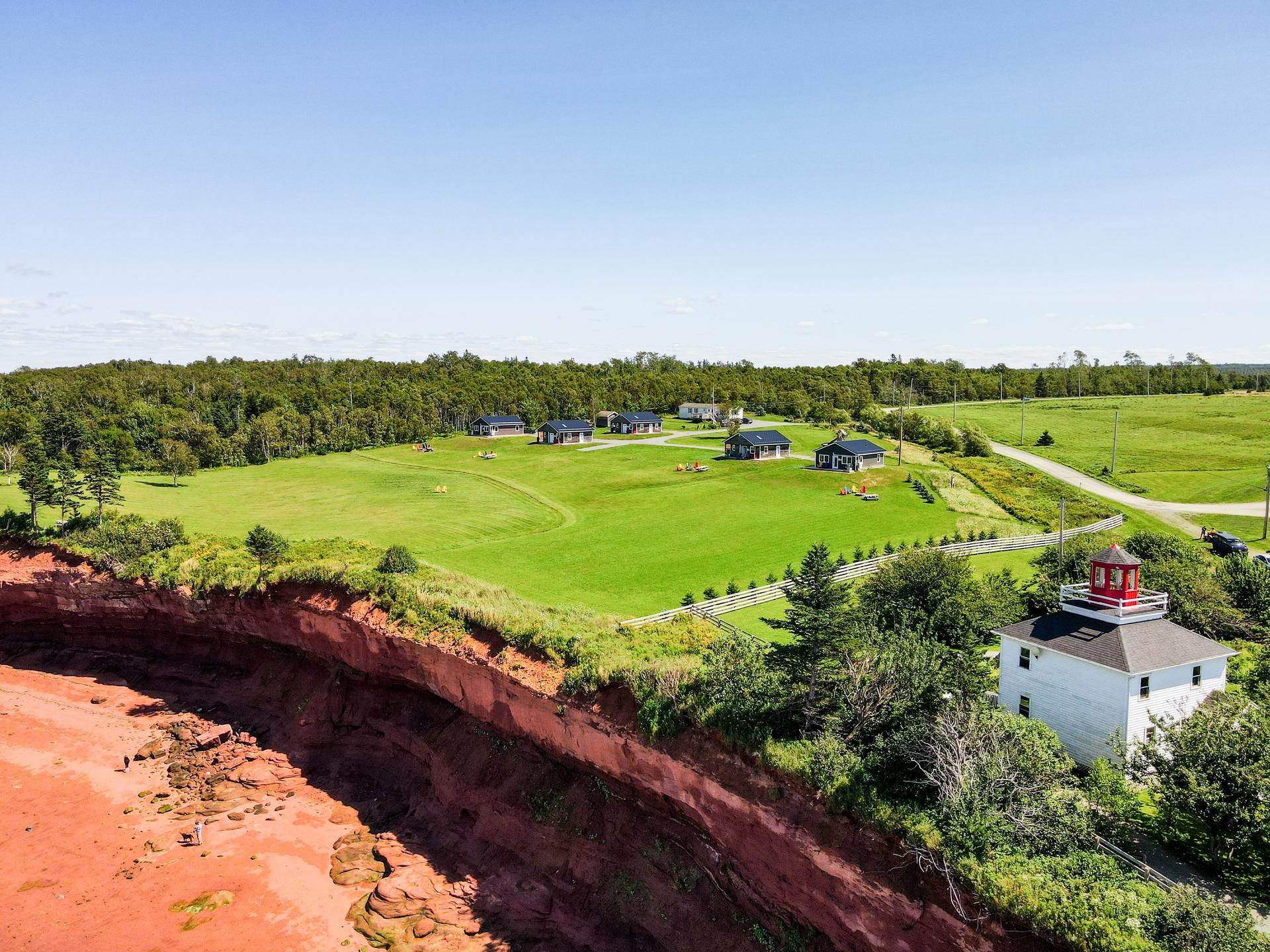 Aerial view of Shangri-La Cottages at Burntcoat Head, Nova Scotia with several cottages on a green clifftop above steep red Bay of Fundy cliffs and a sandy beach, with a small white lighthouse at the cliff's edge.
