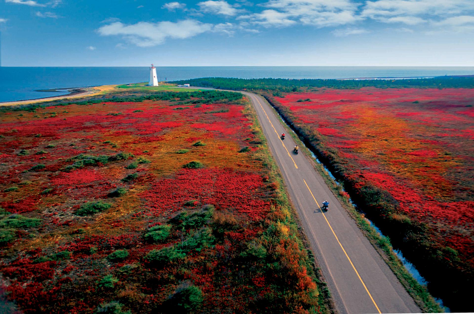 Three people on motorcycles riding towards a lighthouse on the coast of Miscou Island, New Brunswick