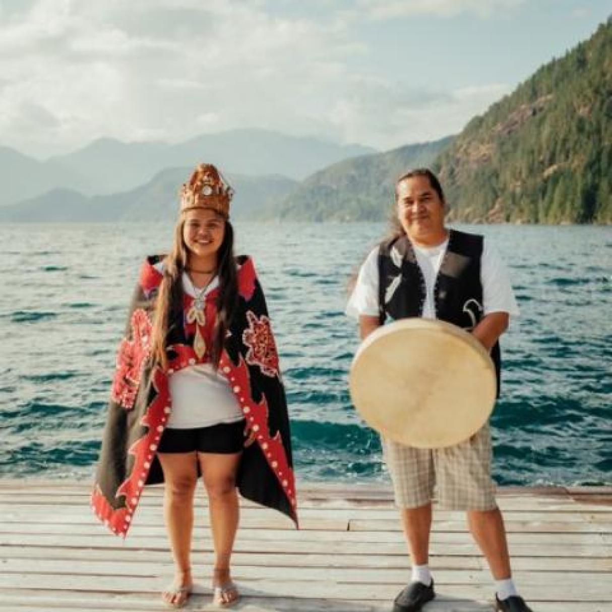 Two people standing on a dock by a lake; one wearing indigenous dress and the other holding an indigenous drum