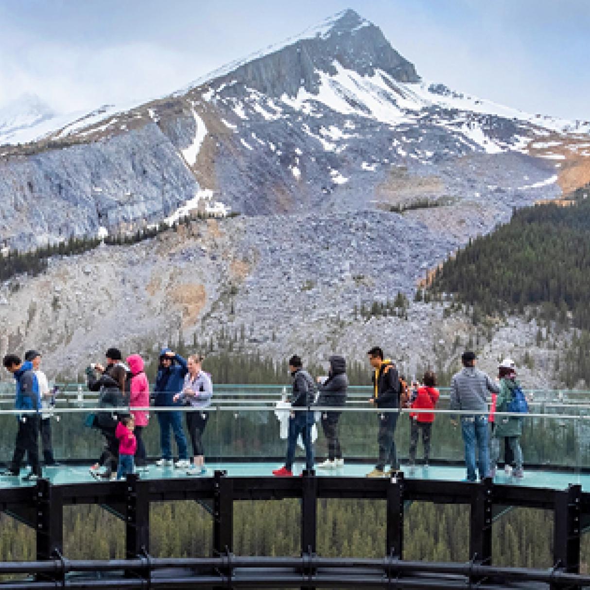 People wstand upon a bridge that overlooks a pointy snocapped mountain. 