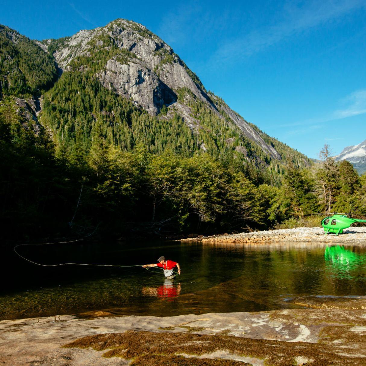 A person stands waist deep and casts a fishing line. In the background sits a green helicopter on the shore accompanied by mountains and forest.