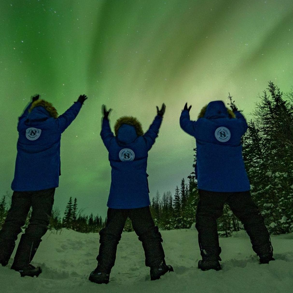 Three people wearing blue parkas hold up their eyes to a green aurora covered night sky.