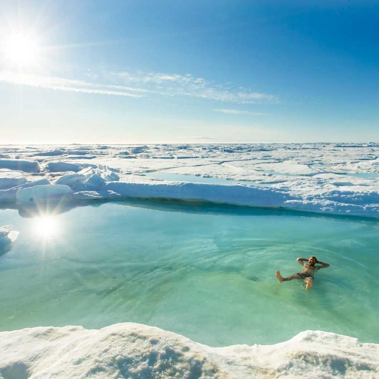 A person with swim shorts sits in a turquoise glacial pool of water surrounded by tundra ice and snow.