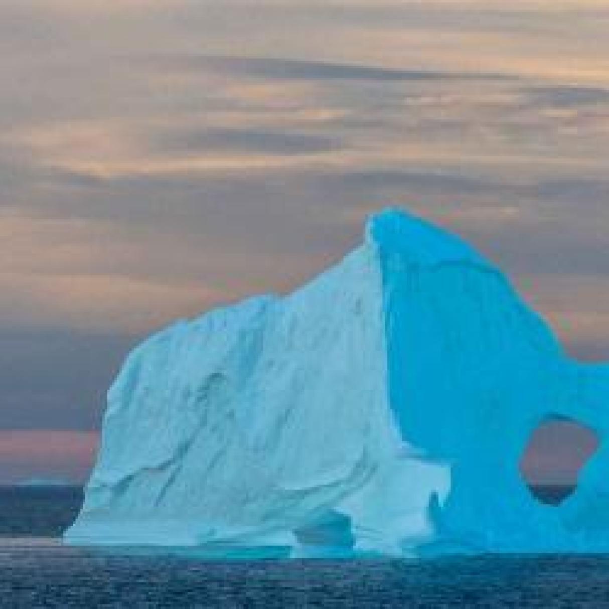 A cool blue iceberg sits in the middle of the ocean in front of a dusk lit sky. 