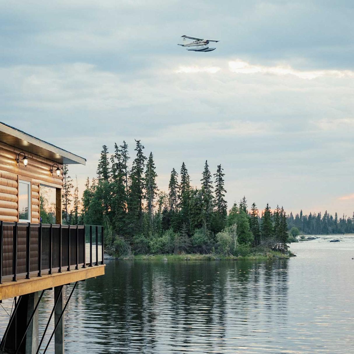 A float plane flys over a forest where a log cabin overlooks a lake