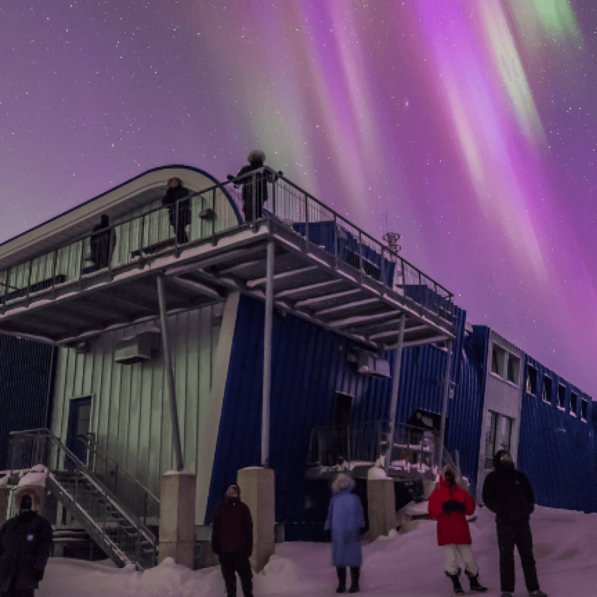 A group of people stand in the snow outside a small building. They look at pink, purple, and green northern lights in the night sky.