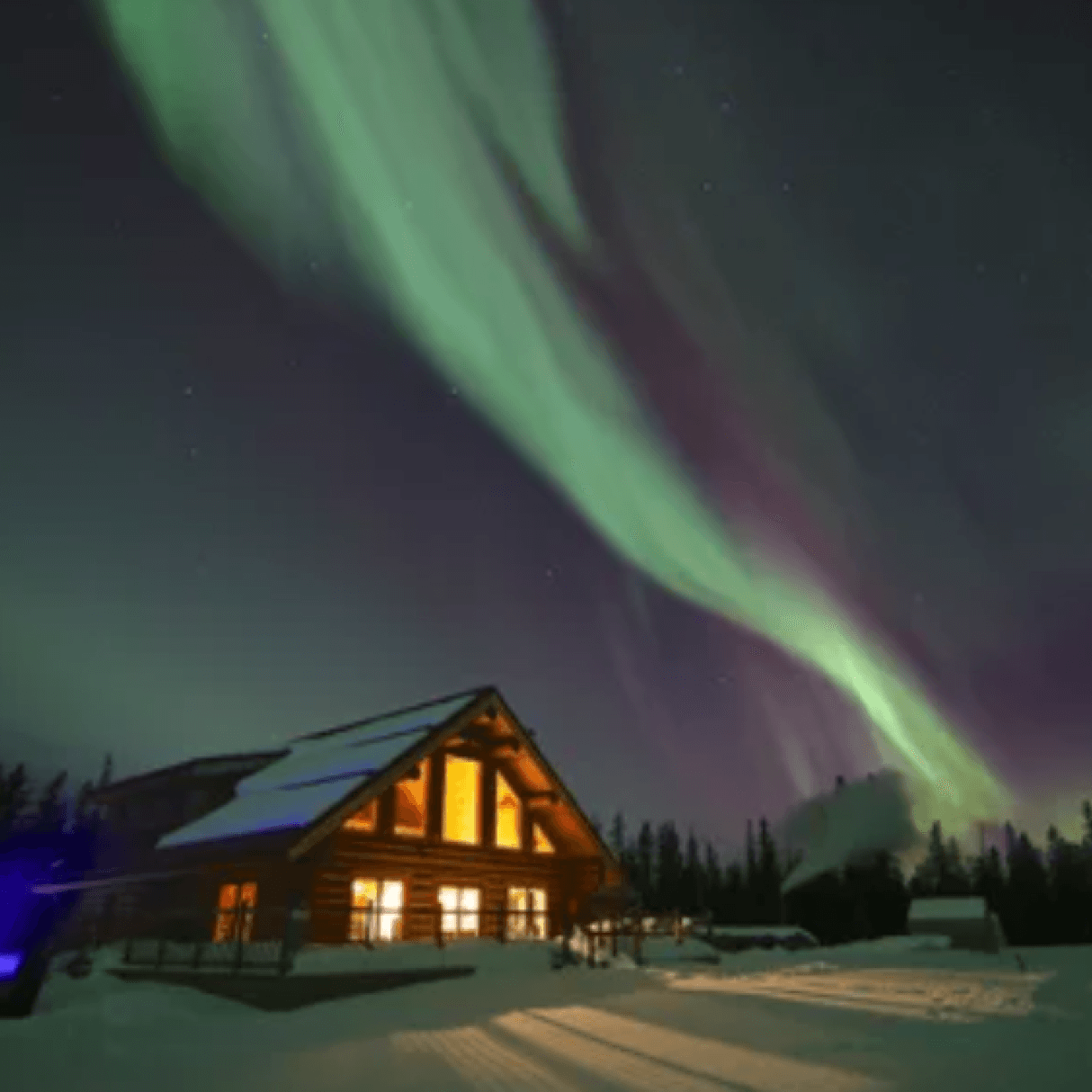 A wooden cabin's windows glow orange in the night. Beside the cabin is a pool. Above, in the night sky, are green northern lights.
