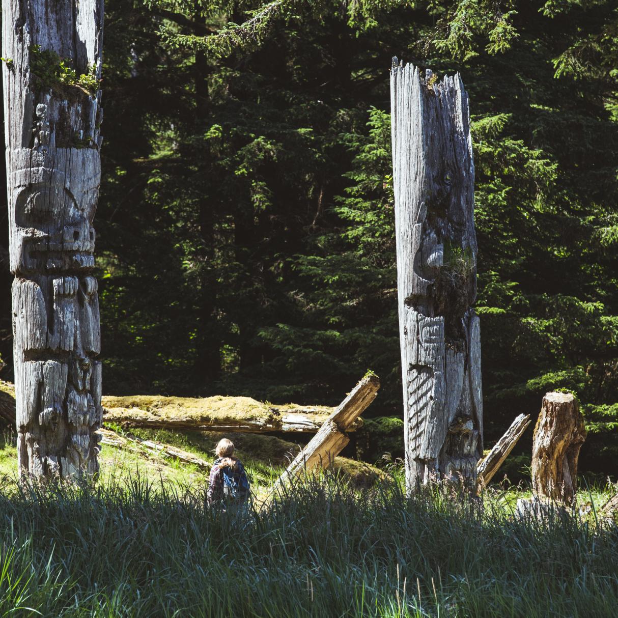 A traveller stands at the base of two totem poles in a forest