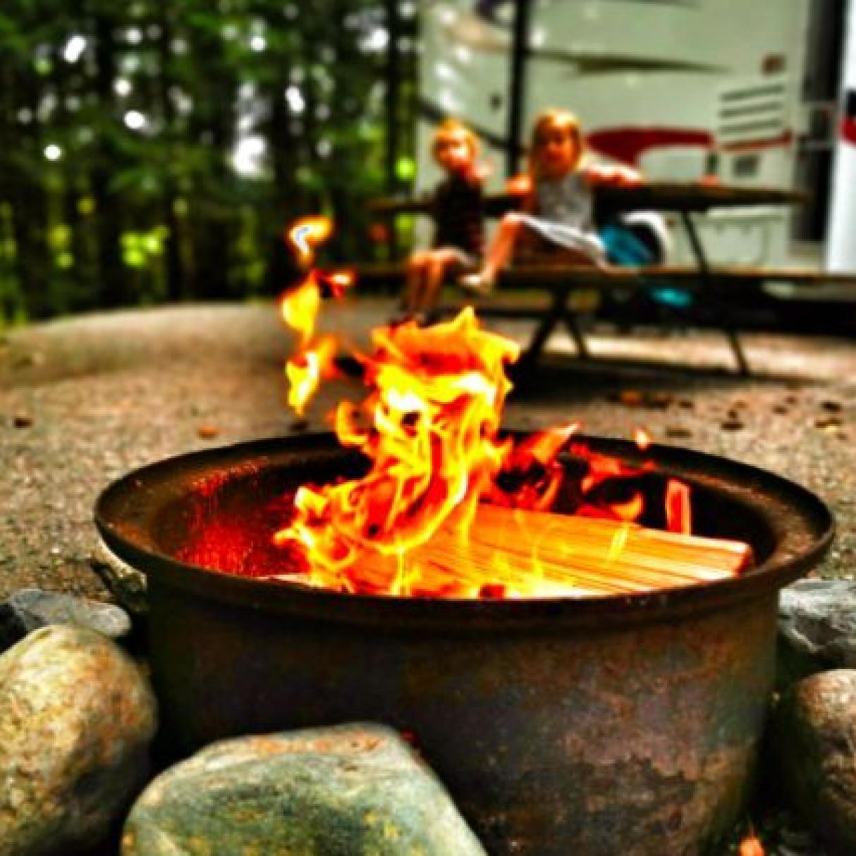A campfire burns with children and an RV in the background 